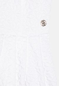 Robe blanche texturée avec un motif géométrique et un design plissé, ornée d'un bouton logo en or et noir.