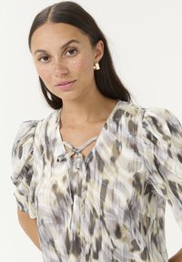 Sheer blouse in soft hues of gray, beige, and white. Features puff sleeves, a V-neck with tie detail, and an abstract pattern throughout.