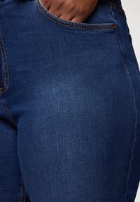 Jeans in denim blu scuro con cuciture arancioni, una tasca frontale e una trama liscia, che evidenziano il peso e la vestibilità del tessuto.