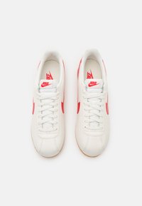 Fehér bőr sportcipők piros részletekkel, texturált felülettel, fűzőzárakkal és gumitalppal. Nike logó a nyelvén és az oldalán.
