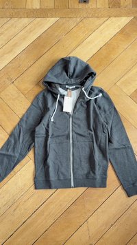 Siva zip-up kapuca s kapuco, z izvlečnimi vrvicami in sprednjimi žepi. Narejena iz mehkega materiala z gladko teksturo in dokončanimi šivi.