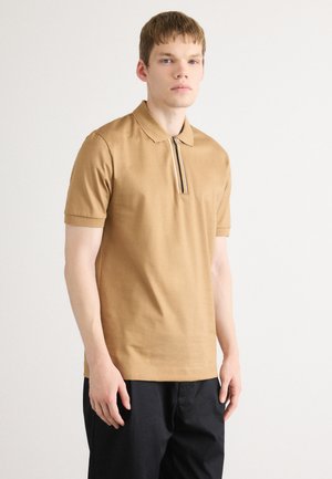 Ung mann står iført en beige kortermet poloskjorte med vertikal glidelås og svarte bukser mot en ensfarget hvit bakgrunn.