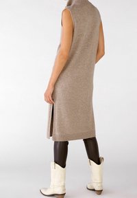 Robe en tricot sans manches, beige, avec un col haut et des fentes sur les côtés, associée à des leggings ajustés marron et des bottes de cowboy blanches.