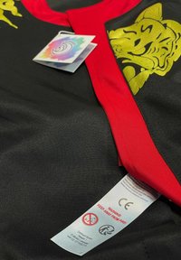 Schwarzes Kleidungsstück mit rotem Kragen und goldenem Tiermotiv. Beinhaltet Etiketten mit Pflegehinweisen und Sicherheitswarnungen.