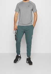 Hombre con camiseta gris de Nike, joggers verdes y zapatillas negras de Nike, sosteniendo una cuerda para saltar negra con la mano izquierda.