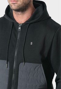 Czarna bluza na zamek z szarym łatką na dolnej części przodu, z kapturem, sznurkami i małym logo na klatce piersiowej.