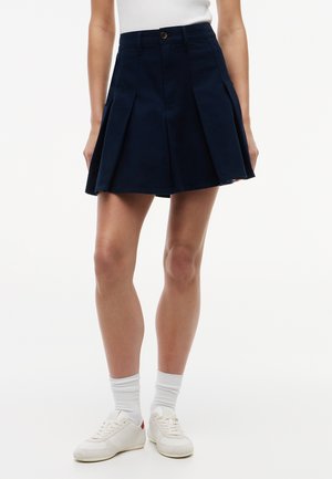 Vrouw draagt een marineblauwe geplooide rok, witte sokken, witte sneakers en een wit mouwloos topje tegen een effen witte achtergrond.