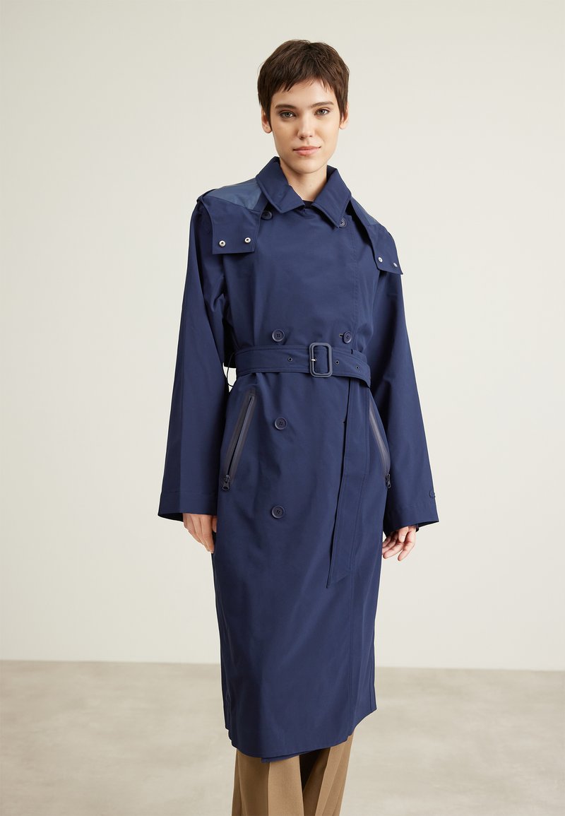 Manteau long bleu marine en tissu lisse. Doté d'une taille cintrée par une ceinture, d'épaulières et de poches zippées. Boutons sur le devant.