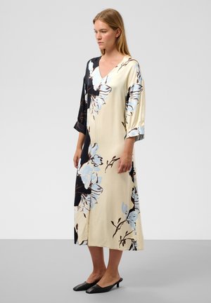 Femme debout portant une longue robe fleurie avec des panneaux noirs et crème, des manches trois-quarts et des chaussures noires à talons bas sur un sol gris.