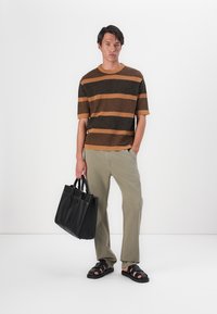 Gestreepte gebreide trui in bruin en oranje, beige broek, zwarte schoudertas en zwarte slipper-sandalen. Man staat voor een witte achtergrond.