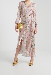 Robe portefeuille florale en tissu rose clair avec manches et ourlet à volants, dotée d'une fente haute. Assortie à un petit sac à main doré et des talons dorés.