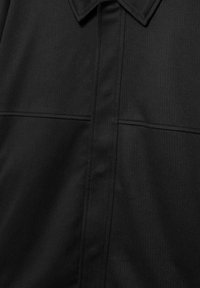 Zwart shirt met een puntige kraag en twee horizontale naaddetails over de borst, stof met subtiele diagonale textuur.