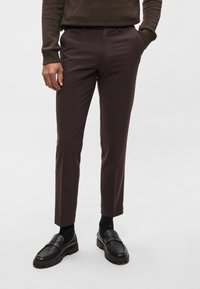 Calças castanhas escuras, de corte ajustado, com frente lisa e bolsos laterais subtilmente definidos. Usadas com mocassins pretos e meias escuras.