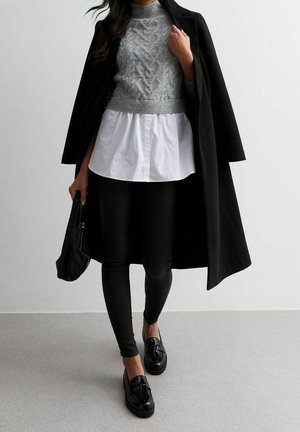 Mujer que lleva un abrigo largo negro sobre un jersey de punto gris y camisa blanca, leggings negros, mocasines negros y sostiene un bolso negro.