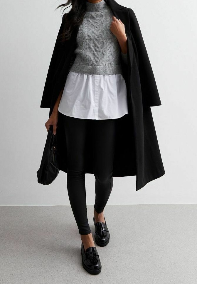 Femme portant un manteau long noir par-dessus un pull en maille gris et une chemise blanche, leggings noirs, mocassins noirs, tenant un sac à main noir.