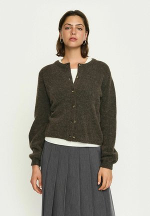 Brown wool cardigan with a round neckline, button front, and puff sleeves. Worn over a white top, paired with a pleated gray skirt.