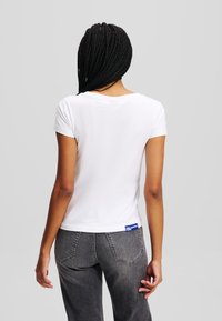 Tricou alb cu maneci scurte și guler rotund, fabricat dintr-un material neted. Spatele prezintă o etichetă albastră aproape de talie, pe blugi negri din denim.
