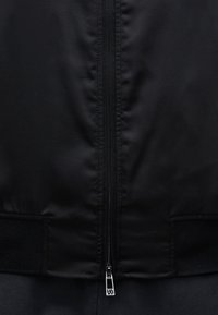 Gros plan de l’avant d’une veste noire montrant une fermeture éclair centrale avec tirette métallique et des poignets en tissu côtelé sur les côtés.