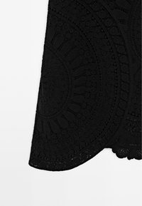 Section de tissu noir avec des motifs en dentelle circulaires et floraux, présentant une broderie complexe et texturée ainsi que des bords festonnés.