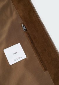 Interior de bolsa de ante marrón con un zipper plateado, que presenta una etiqueta de tela blanca con el nombre de la marca y las instrucciones de cuidado.