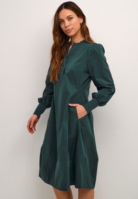 Tannengrünes Kleid aus leichtem Stoff, mit langen Puffärmeln, einem Schalkragen und seitlichen Taschen sowie einem gestuften Saum.