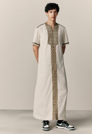 Weißes knielanges Kleidungsstück mit kurzen Ärmeln, das an Halslinie und Vorderseite detaillierte braune geometrische Stickereien aufweist, kombiniert mit schwarzen Turnschuhen.