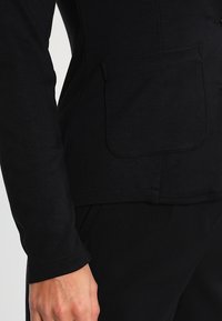 Giacca nera in tessuto testurizzato con tasca laterale, design aderente e maniche lunghe con dettagli di cucitura visibili.
