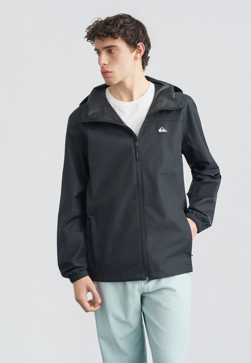 Νεαρός άνδρας με μαύρο φούτερ με φερμουάρ και κουκούλα και ανοιχτό μπλε παντελόνι, κοιτάζει προς τα αριστερά του μπροστά σε μονόχρωμο φόντο.