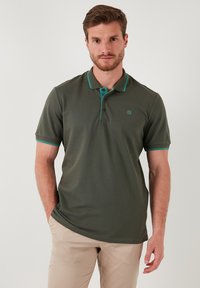 Polo majica s kratkimi rokavi temno zelene barve z teksturirano površino, turkizne poudarke na ovratniku in rokavih ter majhnim logotipom na prsih.