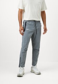 Gaišas, pelēkas sporta bikses ar aukliņu jostā, ar rāvējslēdzēju sānu kabatām un sašaurinātu piegriezumu. Priekšpusē ir zīmola logo.