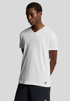 Hvid V-hals T-shirt i bomuldsblanding med korte ærmer og en gul sommerfugle-logo nær kanten, båret med mørke shorts.