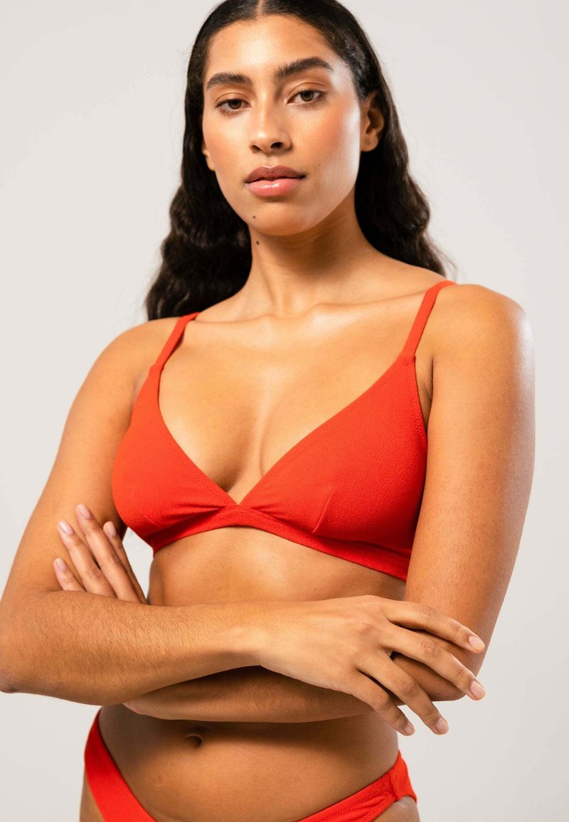 Rode driehoek bikini-top met dunne bandjes. Gelaagde stof, naadloos ontwerp en een enkele twist-detail in het midden. Model poseert met de armen gekruist.