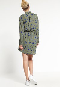 Zelené květinové šaty s modrými květinovými vzory, dlouhými rukávy, stáhnutým pasem a volným střihem. V kombinaci s bílými teniskami.