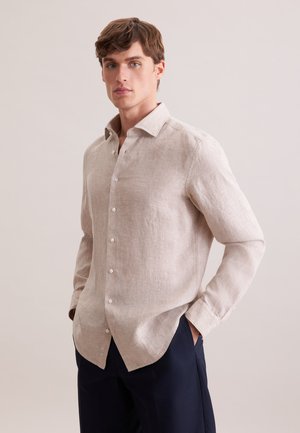 Junger Mann mit hellbeiger, langärmeliger Hemdjacke und dunkler Hose, steht mit den Händen in den Taschen vor neutralem Hintergrund.