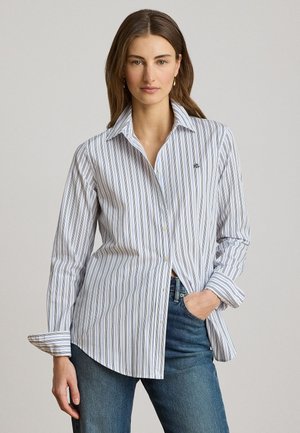 Femme portant une chemise blanche à rayures bleues et un jean, debout avec une main dans la poche devant un fond neutre.