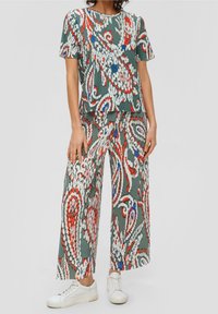 Femme portant un haut à manches courtes assorti et un pantalon large avec un motif floral abstrait coloré, ainsi que des baskets blanches sur un fond gris.