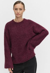Burgundy stickad tröja med en texturerad, fluffig finish; har rund hals och vida ärmar, i kombination med svarta prickiga byxor.