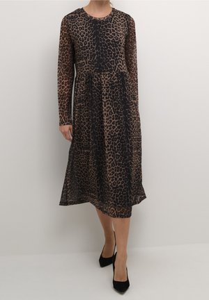 Kobieta ubrana w sukienkę z długim rękawem o długości do połowy łydki w brązowo-czarny wzór cętek lamparta, do tego czarne wysokie szpilki z ostrym noskiem.