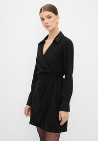 Vestito nero avvolgente con colletto, maniche lunghe e dettaglio alla vita con nastro. Realizzato in un tessuto liscio con un design casual ma strutturato.