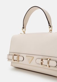 Borsa beige con superficie testurizzata, hardware dorato e tracolle regolabili. Accento triangolare con logo distintivo sul davanti. Forma rettangolare.