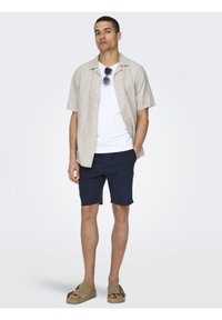 Kortermet beige skjorte med knapper, hvit t-skjorte, marineblå shorts og beige sandaler. Har solbriller og en avslappet passform. Glatt tekstur.