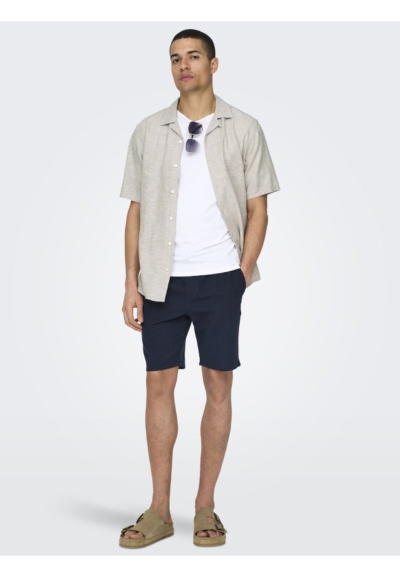 Kortermet beige skjorte med knapper, hvit t-skjorte, marineblå shorts og beige sandaler. Har solbriller og en avslappet passform. Glatt tekstur.