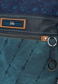 Bolsa de tela azul con estampado, con un accesorio de cuero, que presenta un cierre, una etiqueta marrón con el logotipo "Lois" y un detalle de anillo metálico.