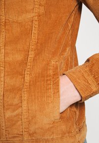Veste en velours côtelé orange brûlé, avec des rainures verticales, une poche poitrine et des manches retroussées. Texture dense avec des accents de coutures visibles.