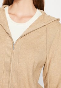 Beżowy hoodie zapinany na zamek wykonany z miękkiego, prążkowanego materiału o lekkiej konstrukcji. Posiada kaptur oraz kontrastującą kremową warstwę wewnętrzną przy dekolcie.