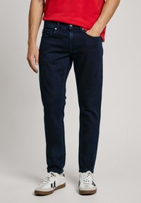 Dark blue jeans with a slim fit, made of denim. Features five pockets and plain design, worn with white and black sneakers.