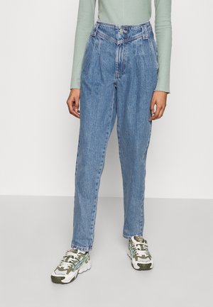 Person wearing high-waisted blue denim jeans, light green long-sleeve top, and white, green, and gray sneakers standing against a plain background.