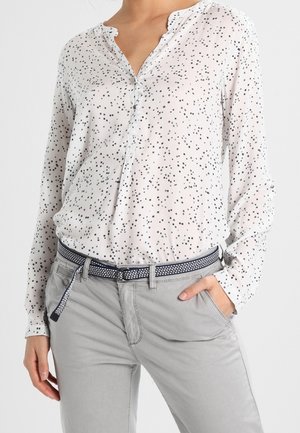 Woman wearing a white long-sleeve blouse with black star prints, tucked into light grey pants with a black and white patterned belt.