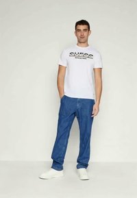 Man standing wearing white Guess t-shirt, blue jeans, and white sneakers against a plain light background.
