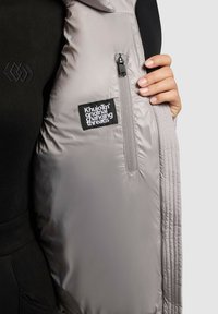 Giubbotto puffer grigio chiaro con una texture trapuntata, dotato di una zip laterale e un'etichetta nera con scritto "Khujo Original Changing Threads."